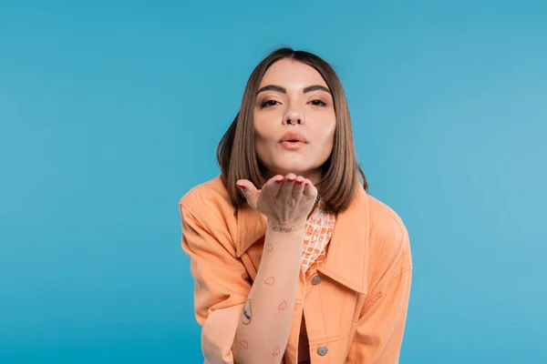 Sending air kiss, brunette young woman with short hair, piercing in nose and tattoos posing in casual outfit on blue background, everyday makeup, orange shirt, generation z, blow kiss — Stock Photo