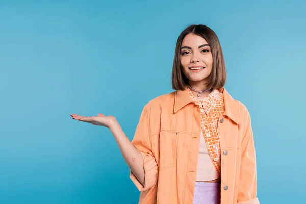 Happy young woman with short hair, piercing in nose and tattoos demonstrating something in camera on blue background, showing, orange shirt, summer outfit generation z — Stock Photo