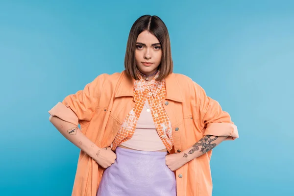 Offended young woman standing with hands on hips on blue background, looking at camera, summer outfit, emotional, displeased, generation z, short brunette hair, pierced nose, tattooed — Stock Photo