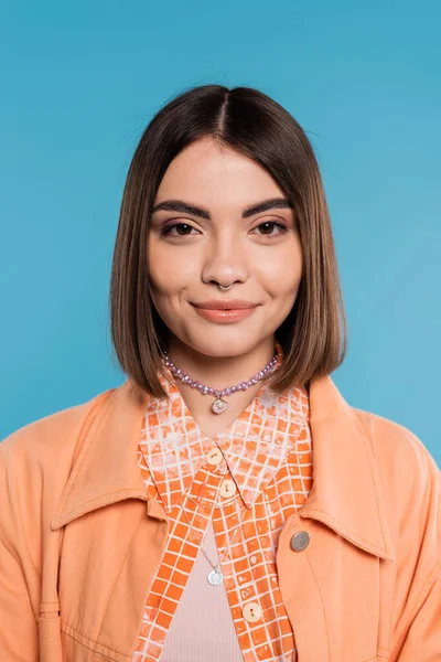 Portrait of cheerful woman, young fashion model smiling and looking at camera on blue background, orange shirt, generation z, short brunette hair, pierced nose, summer outfit, gen z fashion — Stock Photo