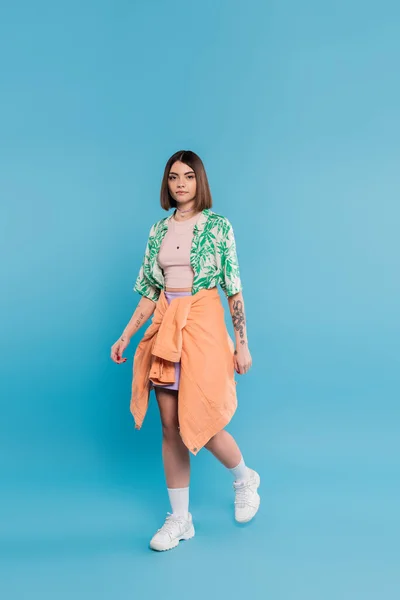 Everyday style, young brunette woman with short hair walking in shirt with palm tree print, skirt and white sneakers on blue background, carefree, tattooed, nose piercing, casual attire, full length — Stock Photo