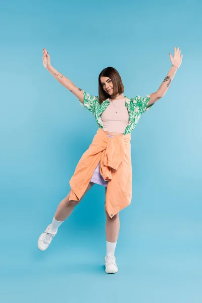 Gen z fashion, carefree woman with short hair posing in shirt with palm tree print, skirt and white sneakers on blue background, tattooed, nose piercing, casual attire, raised hands, full length — Stock Photo