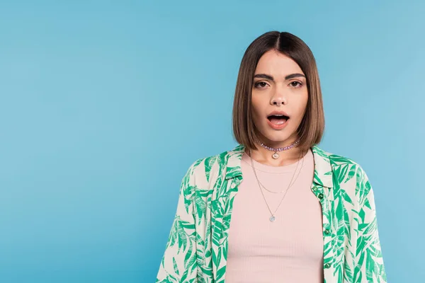 Mujer emocional con la boca abierta, modelo sorprendido mirando a la cámara sobre fondo azul, camisa con impresión de palmera, piercing en la nariz, atuendo casual, generación z, cara sorprendida - foto de stock