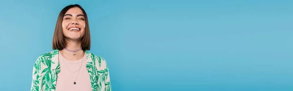 Cheerful young woman with short brunette hair wearing shirt with palm tree print, smiling with closed eyes on blue background, casual attire, gen z fashion, emotional, happiness, banner — Stock Photo