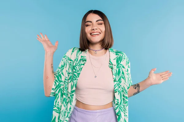 Tattooed young woman with short brunette hair wearing shirt with palm tree print, smiling with closed eyes and gesturing with hands on blue background, casual attire, gen z fashion, happiness — Stock Photo