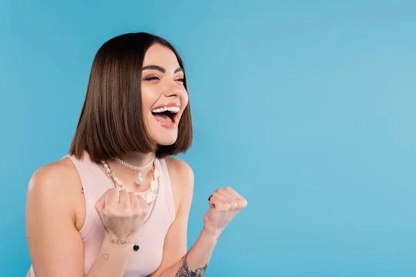 Excitement, tattooed young woman with short brunette hair in tank top smiling and gesturing with hands on blue background, casual attire, gen z fashion, happiness — Stock Photo