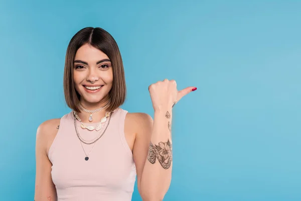 Cheerfulness, tattooed young woman with short brunette hair in tank top smiling and pointing with thumb on blue background, casual attire, gen z fashion, happiness, showing something on camera — Stock Photo