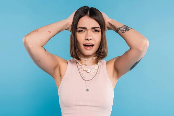 Failure, upset young woman with short brunette hair and tattoos holding hands near head and looking at camera on blue background, casual attire, gen z fashion, emotional — Stock Photo