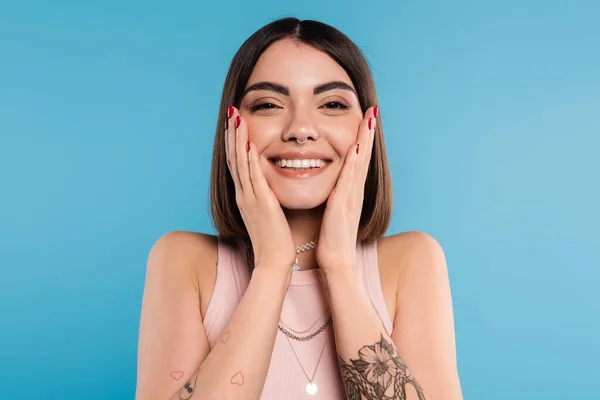 Positivity, tattooed young woman with short brunette hair in tank top smiling  and touching her face on blue background, casual attire, gen z fashion, happiness, joyful face — Stock Photo