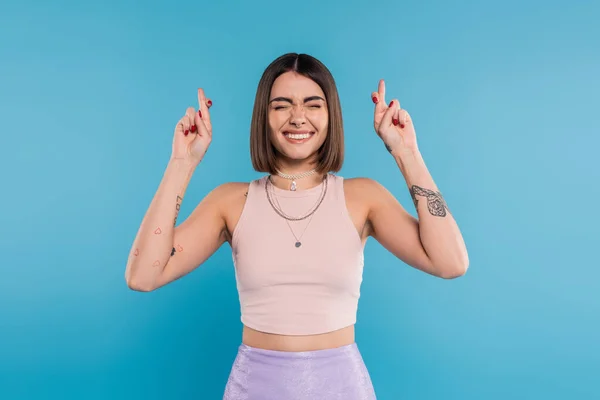Fingers crossed, tattooed young woman with short brunette hair in tank top making wish on blue background, casual attire, gen z fashion, happiness, smiling with closed eyes — Stock Photo