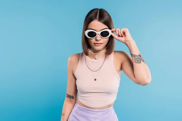 Gen z fashion, brunette young woman with short hair in tank top, skirt and sunglasses posing on blue background, casual attire, stylish posing, personal style, portrait, fashionista — Stock Photo