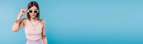 Mode moderne, jeune femme brune aux cheveux courts en débardeur, jupe et lunettes de soleil posant sur fond bleu, tenue décontractée, pose élégante, style personnel, bannière — Photo de stock