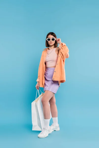 Trendy shopping, brunette young woman in tank top, skirt, sunglasses and orange shirt posing with shopping bag on blue background, casual attire, stylish posing, gen z, modern fashion — Stock Photo