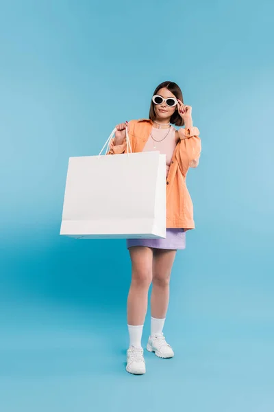 Einkaufsbummel, brünette junge Frau im Tank-Top, Rock, Sonnenbrille und orangefarbenem Hemd posiert mit Einkaufstasche auf blauem Hintergrund, lässige Kleidung, stilvolles Posen, moderne Mode — Stockfoto