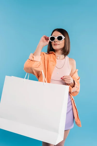 Летние шоппинг, модная молодая женщина в майке танка, юбка, солнцезащитные очки и оранжевая рубашка позирует с сумкой на синем фоне, случайный наряд, стильная поза, ген z, современная мода — стоковое фото