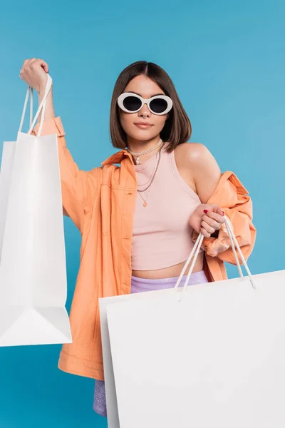 Sommereinkauf, Generation Z, brünette junge Frau im Tank-Top, Rock, Sonnenbrille und orangefarbenem Hemd posiert mit Einkaufstaschen auf blauem Hintergrund, lässige Kleidung, stilvolles Posen, moderne Mode — Stockfoto