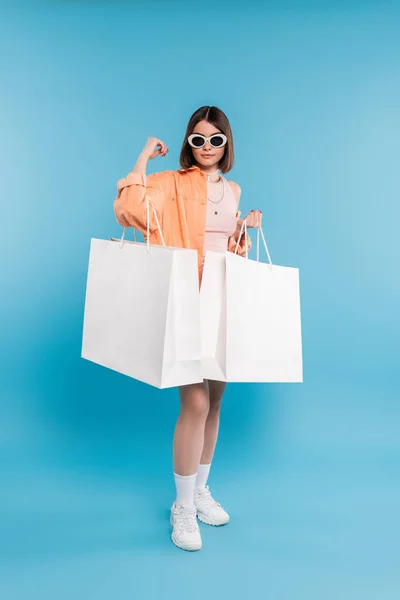 Sommereinkauf, Modetrend, brünette junge Frau in Tank Top, Rock, Sonnenbrille und orangefarbenem Hemd posiert mit Einkaufstaschen auf blauem Hintergrund, stylisches Posing, Gen Z, moderne Mode, volle Länge — Stockfoto