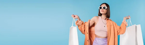 Sommerspaß, brünette junge Frau mit Sonnenbrille und trendigem Outfit posiert mit Einkaufstaschen auf blauem Hintergrund, lässige Kleidung, stilvolles Posing, Generation Z, Modetrend, Banner — Stockfoto