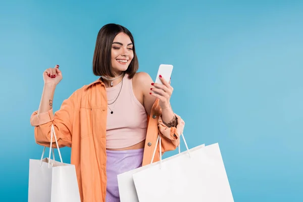 Позитивность, шоппинг, веселая молодая женщина в модном наряде держа сумки и используя смартфон на синем фоне, случайный наряд, стильный, поколение z, современная мода — стоковое фото