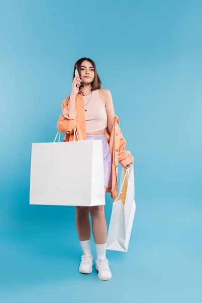 Позитивность, шоппинг, телефонный звонок, веселая молодая женщина в модном наряде держа сумки и разговаривая на смартфоне на голубом фоне, случайный наряд, поколение z, современная мода — стоковое фото
