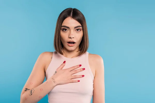 Shocked woman with hand on chest, stylish model with tattoos and nose piercing looking at camera on blue background, emotional, opened mouth, generation z, summer fashion — Stock Photo