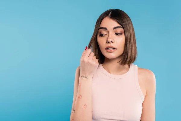 Hair damage, stress, concerned brunette young woman touching her hair on blue background, tattooed, nose piercing, tank top, generation z, casual attire, thinning hair — Stock Photo