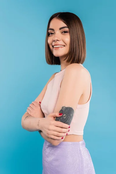 Folded hands, cheerful young woman with short hair and tattoos standing and looking at camera on blue background, nose piercing, youth, generation z, summer fashion — Stock Photo