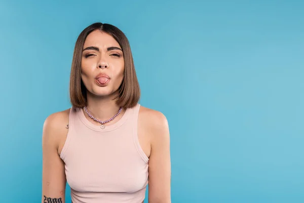 Tattooed young woman with nose piercing and short hair sticking out tongue on blue background, generation z, fashionable model, trendy summer fashion, youth culture — Stock Photo