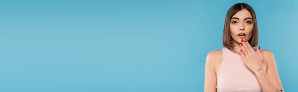 Espantado, tatuado jovem mulher com nariz piercing e cabelo curto segurando mão perto da boca no fundo azul, geração z, moda, moda de verão na moda, banner — Fotografia de Stock