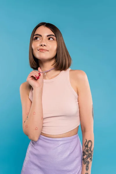 Dreamy, tattooed young woman with nose piercing and short hair touching silver necklace and looking away on blue background, generation z, fashionable, trendy summer fashion — Stock Photo
