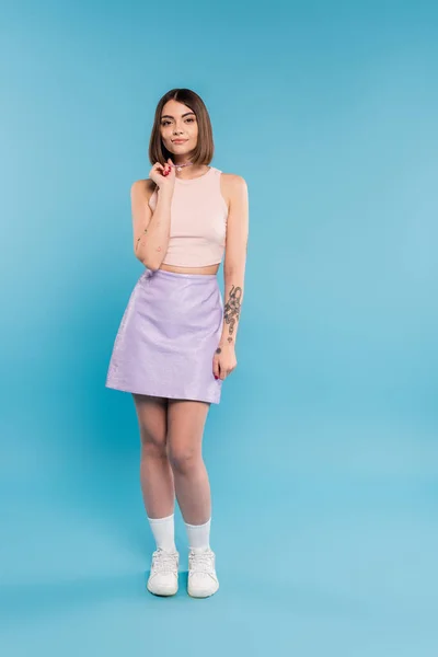 Fashion trend, brunette young woman with short hair in tank top, skirt and white sneakers posing on blue background, casual attire, gen z fashion, personal style, nose piercing, everyday makeup — Stock Photo