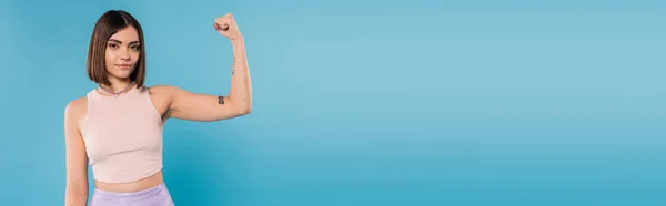 Frauenpower, brünette junge Frau mit kurzen Haaren, Tätowierungen und Nasenpiercing, die Muskeln auf blauem Hintergrund zeigt, Generation z, unzufrieden, Freizeitkleidung, Stärke, Banner — Stockfoto