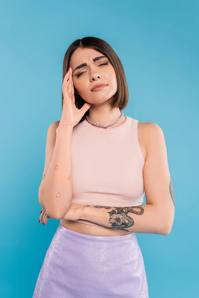 Having headache, brunette young woman with short hair, tattoos and nose piercing touching head on blue background, generation z, displeased, casual attire, migraine, feeling pain — Stock Photo