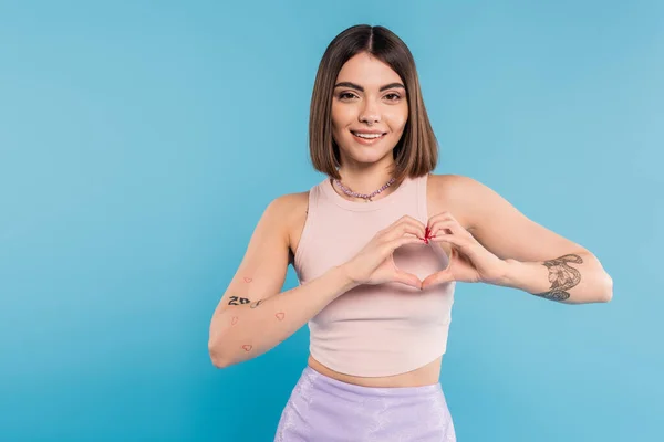 Positivity and love, happy young woman with short hair, tattoos and nose piercing showing heart gesture with hands on blue background, generation z, cheerful, casual attire — Stock Photo