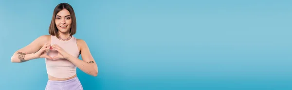 Positivität und Liebe, glückliche junge Frau mit kurzen Haaren, Tätowierungen und Nasenpiercing, die Herzgeste mit Händen auf blauem Hintergrund zeigt, Generation z, fröhlich, lässig gekleidet, Banner — Stockfoto