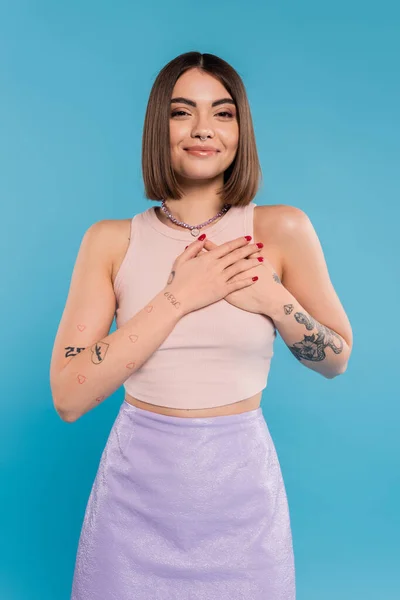 Positivity and peace, happy young woman with short hair, tattoos and nose piercing holding hands near chest on blue background, generation z, cheerful, casual attire — Stock Photo