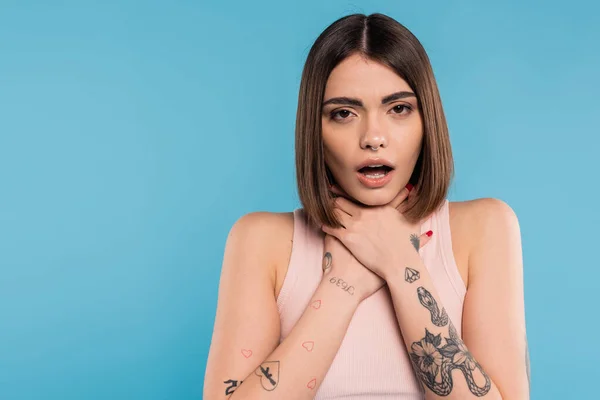 Brunette young woman with short hair holding hands near neck and standing with opened mouth while choking on blue background, casual attire, gen z fashion, personal style, nose piercing — Stock Photo