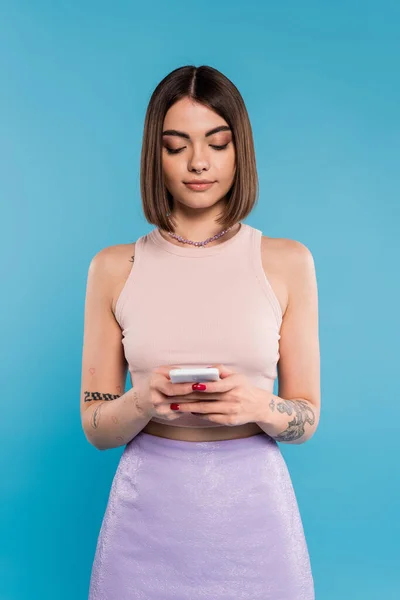 Mujer mensajes de texto en el teléfono inteligente, pelo corto, tatuajes y piercing en la nariz usando el teléfono móvil sobre fondo azul, atuendo casual, moda gen z, estilo personal, maquillaje diario - foto de stock