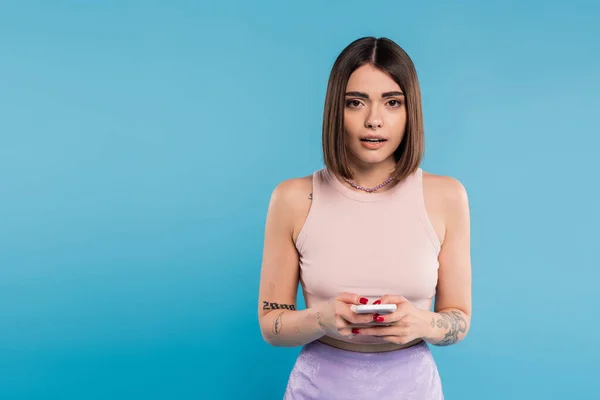 Messaging on smartphone, shocked, young brunette woman short hair, tattoos and nose piercing using mobile phone on blue background, casual attire, gen z fashion, social media influencers — Stock Photo