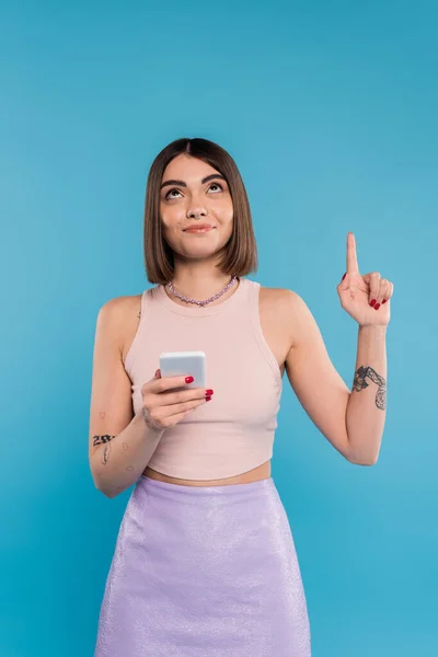 Holding smartphone, young brunette woman short hair, tattoos and nose piercing pointing up on blue background, casual attire, gen z fashion, social media influencers — Stock Photo