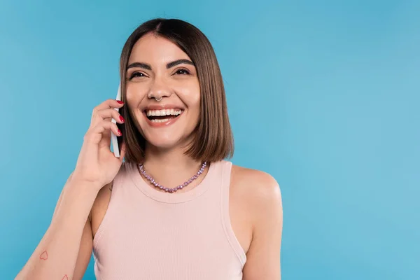Phone call, positivity, cheerful young woman with short hair, tattoos and nose piercing talking on smartphone on blue background, casual attire, gen z fashion, personal style — Stock Photo