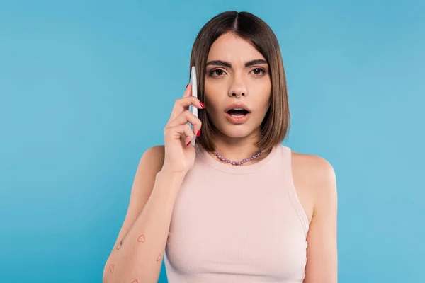 Phone call, shocked young woman with short hair, tattoos and nose piercing talking on smartphone on blue background, casual attire, gen z fashion, opened mouth, emotional — Stock Photo