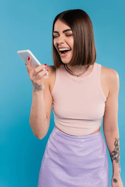 Botschaften senden, aufgeregte junge Frau kurze Haare, Tätowierungen und Nasenpiercing mit dem Handy auf blauem Hintergrund, lässige Kleidung, Gen-Z-Mode, Social-Media-Influencer — Stockfoto