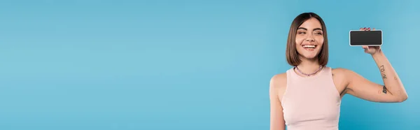 Smartphone mit leerem Bildschirm, attraktive junge Frau mit kurzen Haaren, Tattoos und Nasenpiercing, Handy auf blauem Hintergrund, Gen-Z-Mode, Social-Media-Influencer, Banner — Stockfoto