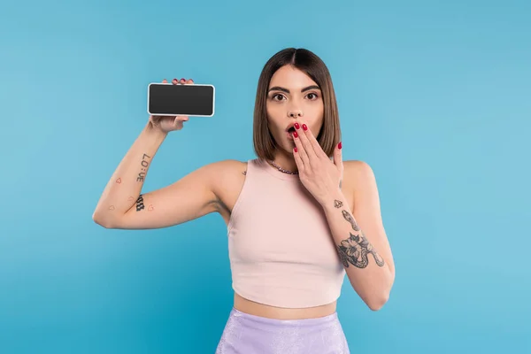 Смартфон с чистым экраном, шокированная молодая женщина с короткими волосами, татуировки и пирсинг носа держа мобильный телефон на синем фоне, ген z мода, социальные медиа влиятельные, прикрыть рот — стоковое фото