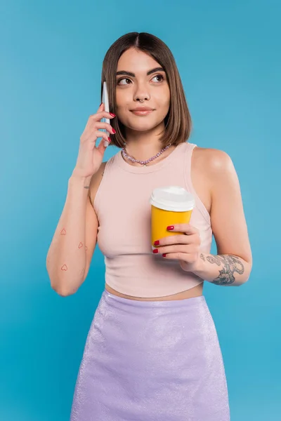 To go coffee, happy and tattooed young woman with short hair and nose piercing holding paper cup and talking on smartphone on blue background, generation z, summer trends, attractive, coffee culture — Stock Photo