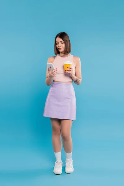 To go coffee, tattooed young woman with short hair and nose piercing holding paper cup and talking on smartphone on blue background, generation z, summer trends, attractive, full length — Stock Photo