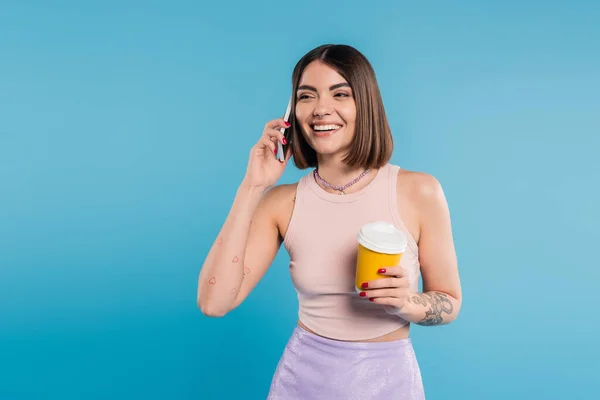 Zum Kaffeetrinken, fröhliche und tätowierte junge Frau mit kurzen Haaren und Nasenpiercing, Pappbecher in der Hand und Smartphone auf blauem Hintergrund, Generation Z, attraktiv, Kaffeekultur — Stockfoto