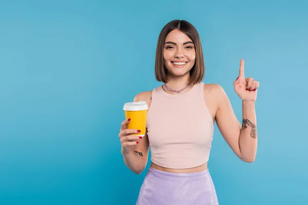 Para ir café, mujer joven feliz con el pelo corto, tatuajes y piercing en la nariz sosteniendo taza de papel sobre fondo azul, generación z, tendencias de verano, atractivo, cultura del café, estilo cotidiano, apuntando hacia arriba - foto de stock