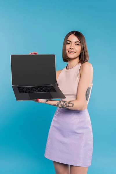Freelancer, cheerful young woman with short hair, tattoos and nose piercing holding laptop on blue background, generation z, summer trends, attractive, remote work, everyday style, blank screen — Stock Photo